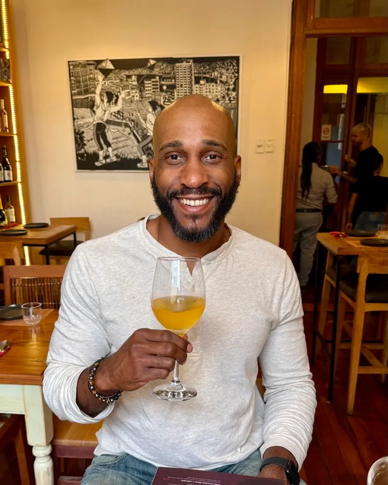 Manuel Patterson smiling and holding a glass of orange wine at a restaurant table