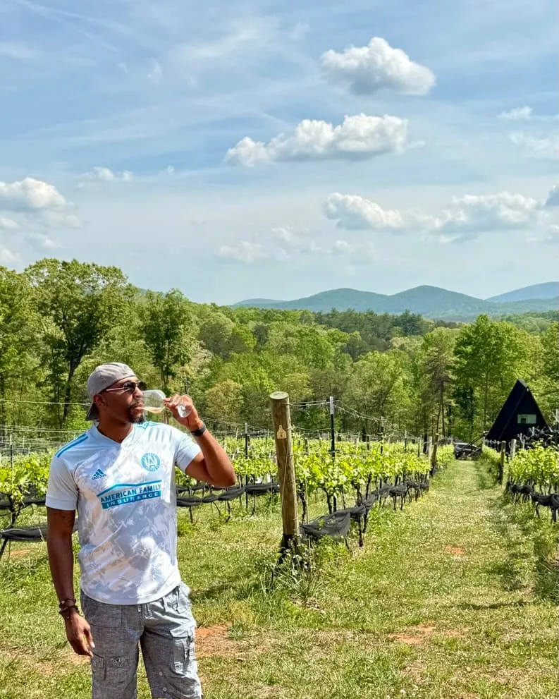 Manuel Patterson sipping a glass of white wine between vineyard rows with mountains behind